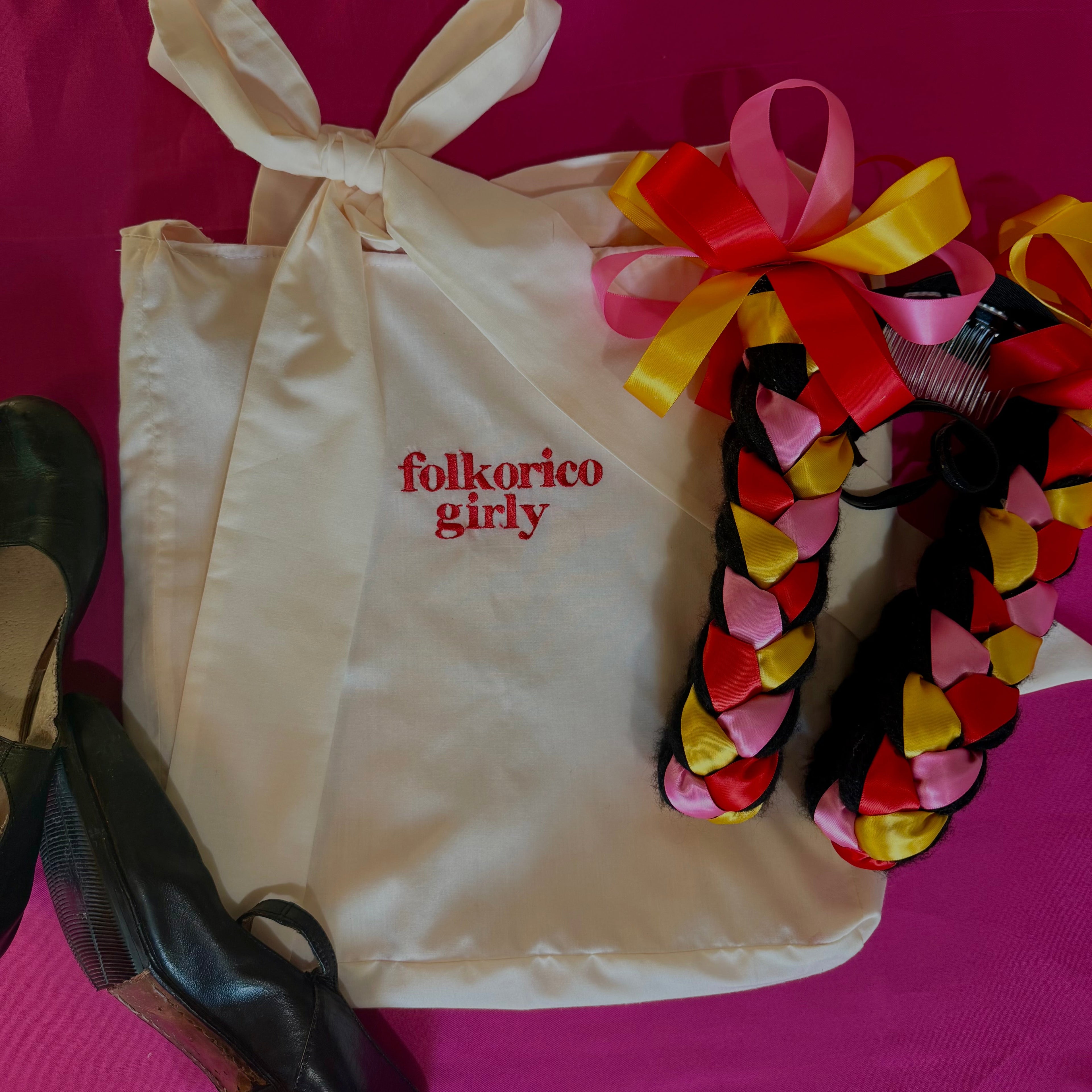 Beige/off white tote bag with 'folklorico girly' embroidered text, colorful braided hair accessory with ribbons, and black folklorico shoes on a pink background