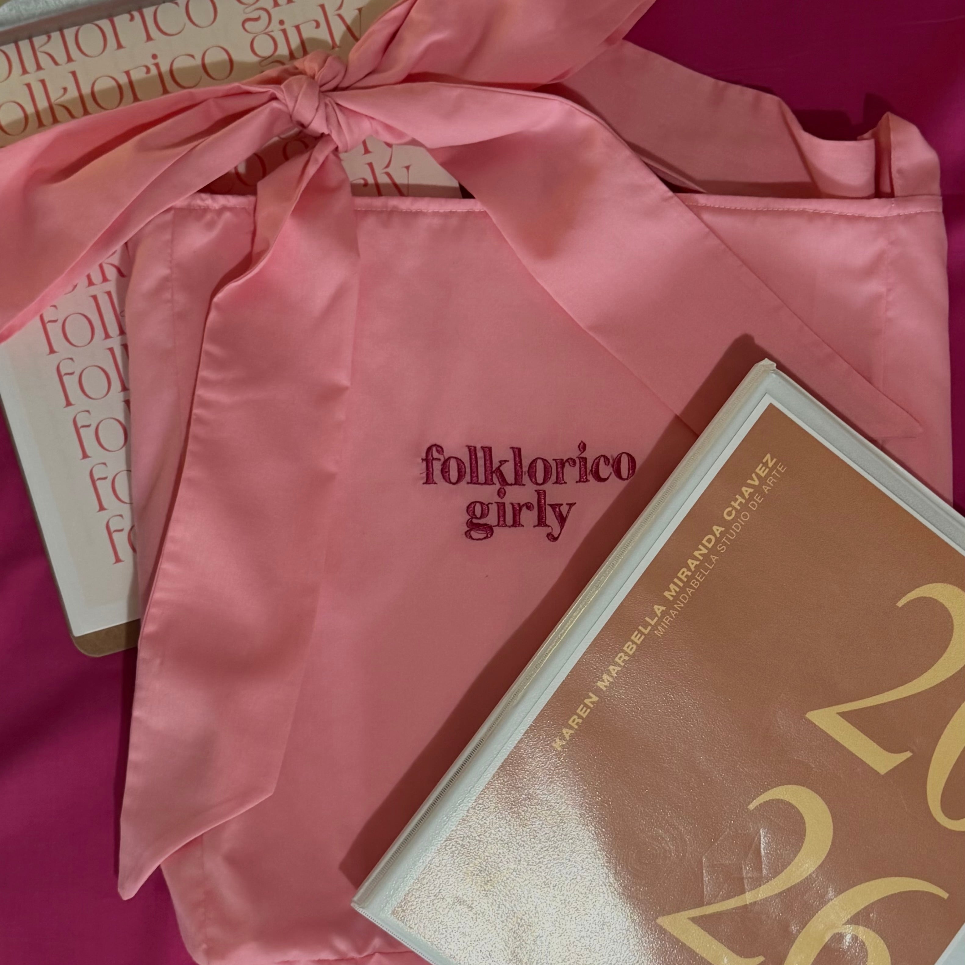 Pink tote bag with 'folklorico girly' embroidered text next to a binder and clipboard, on a pink background