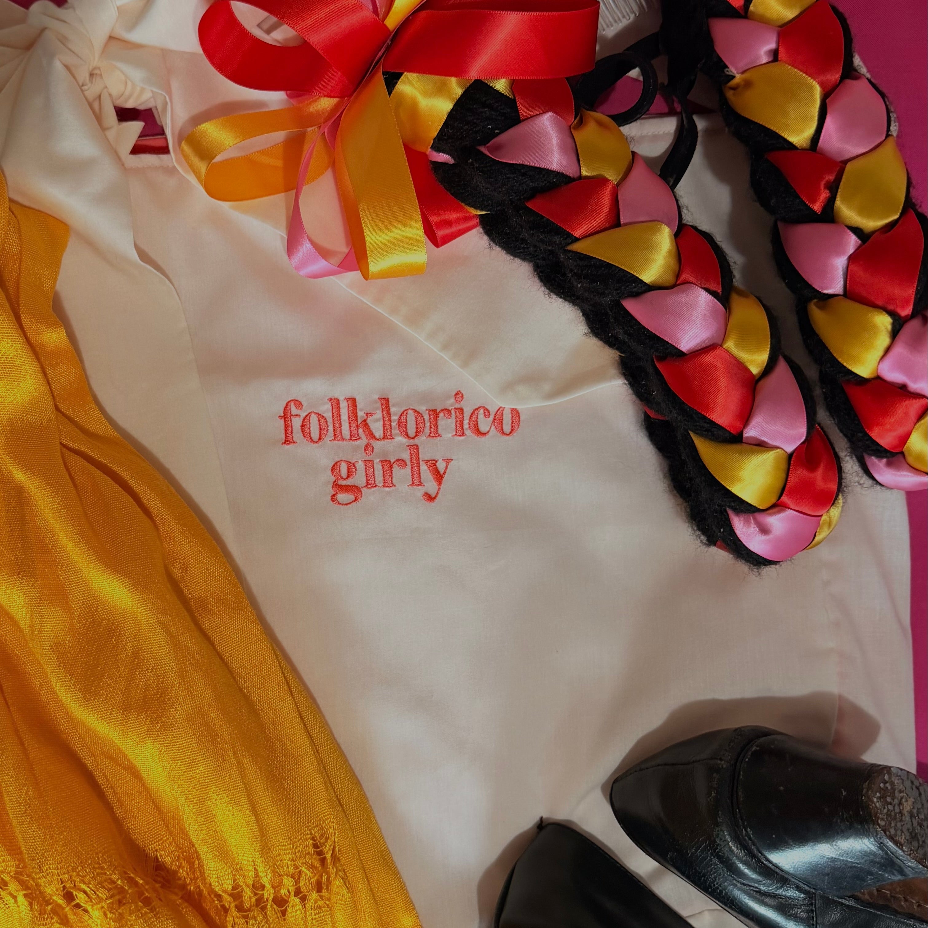 Beige, off white tote bag with coral Embroidered “folklorico girly” text, next to colorful ribbons on a braided hair piece and some folklorico heels. 