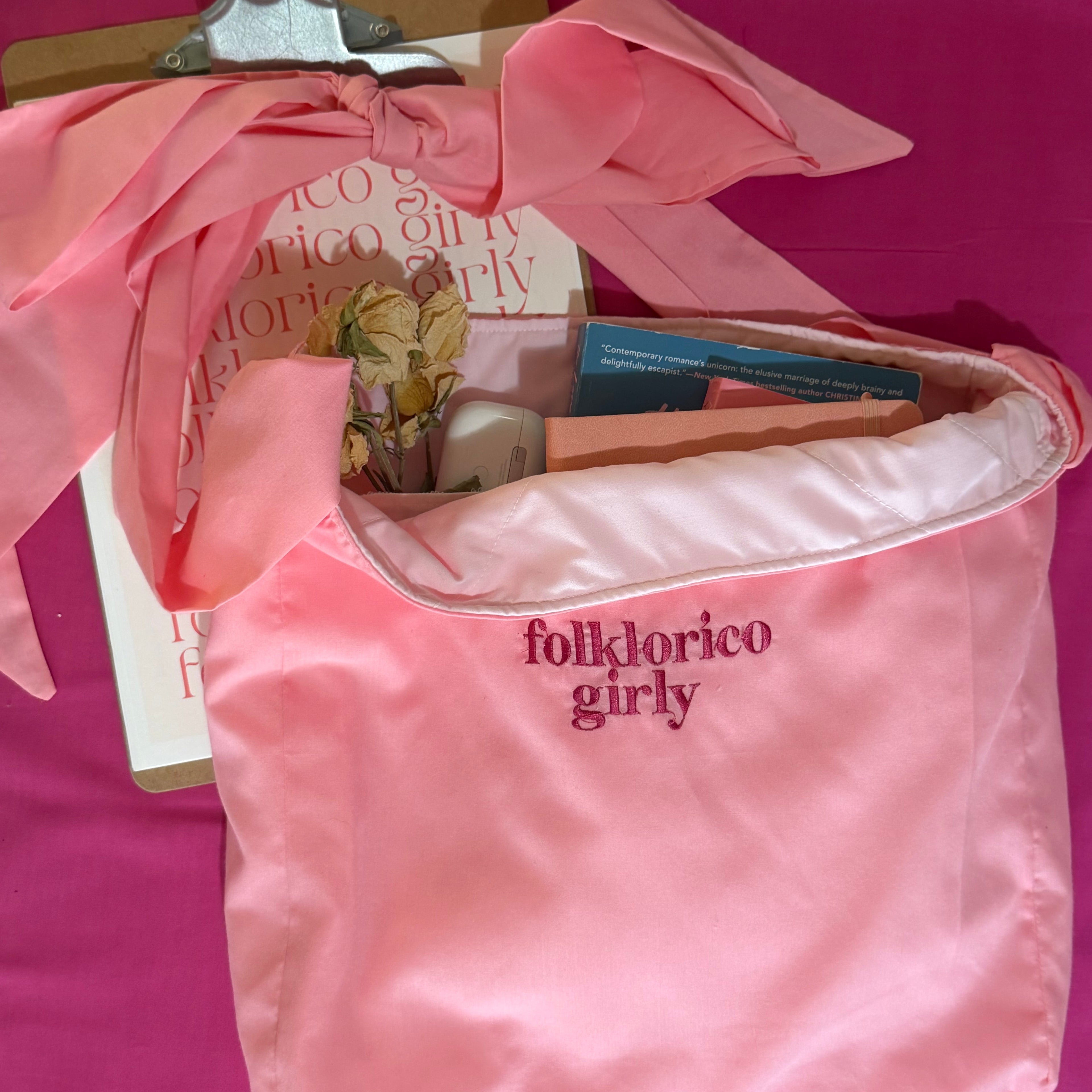 Pink tote bag  stuffed with books and small trinkets, with magenta, embroidered “folklorico girly” branding on a pink background