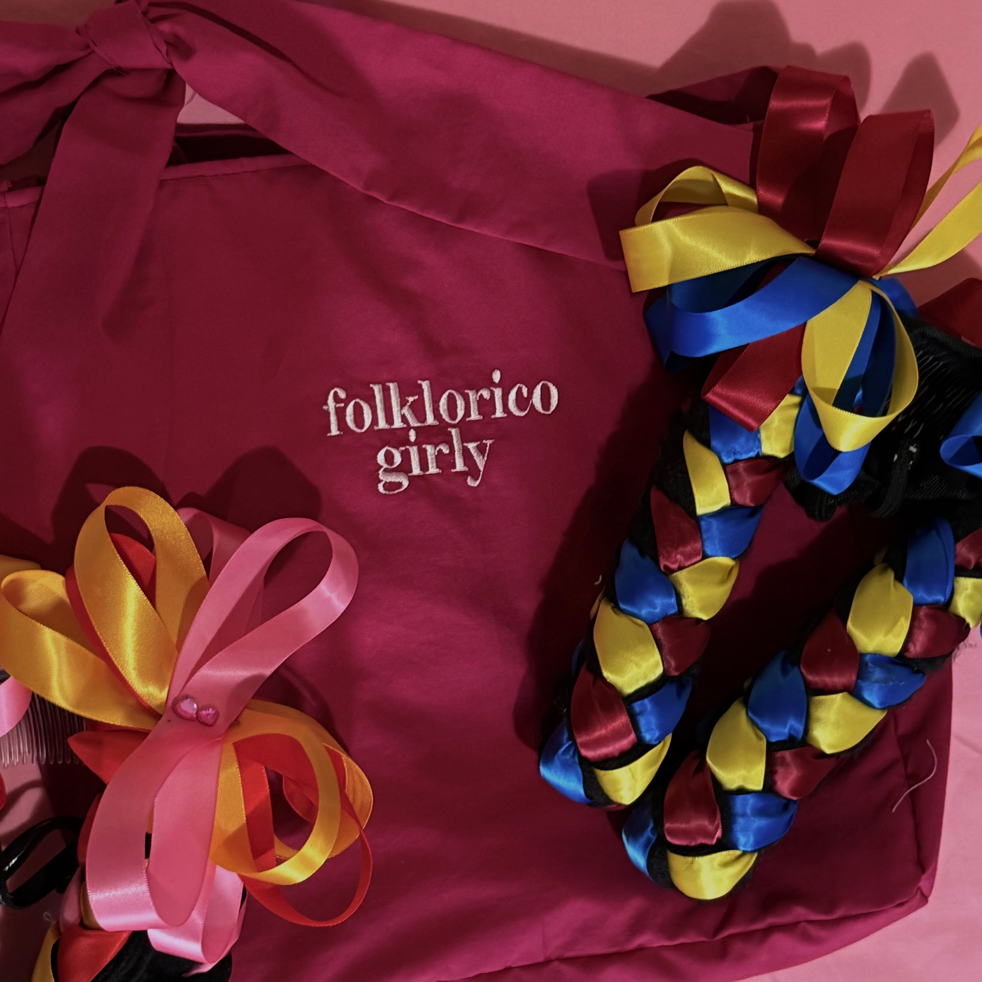 Colorful braided hair accessory with ribbons on top of a magenta tote bag featuring white, embroidered 'folklorico girly' text.